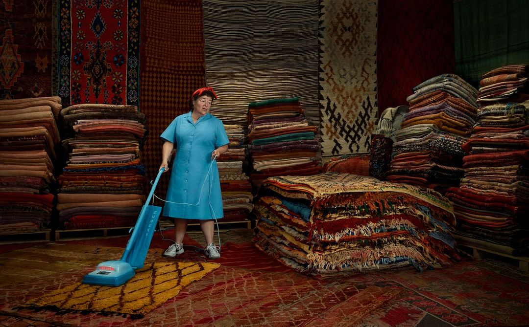 Mature woman in a blue dress vacuums a patterned rug surrounded by tall stacks of colorful rugs and carpets in a large warehouse setting.